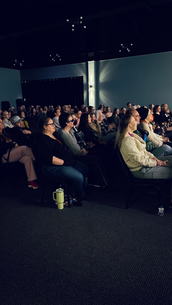The people seated in the auditorium are viewed from the side. The audience's attention is directed towards an unseen proje...