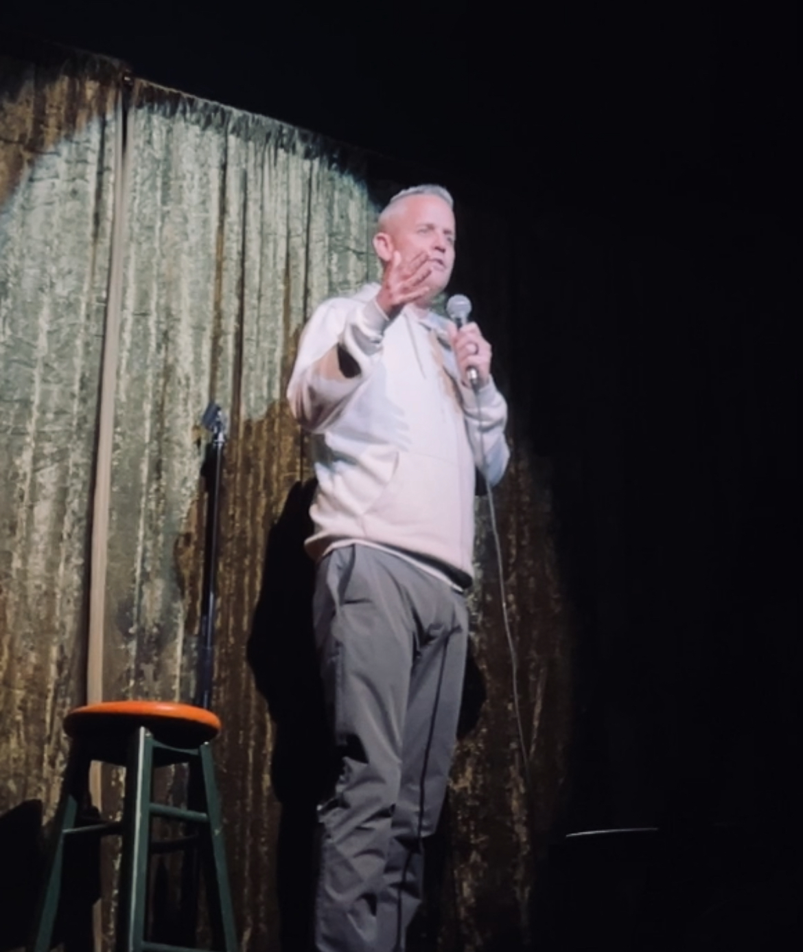 A man is illuminated on stage, stood behind a microphone, although the image is blurry. He wears a white hoodie and grey p...