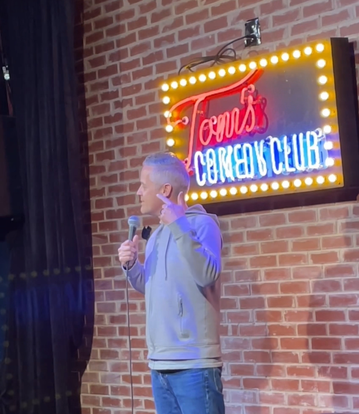 Here is the alt text for the image: "A man in a light hoodie and jeans stands on stage, speaking into a microphone, with a...