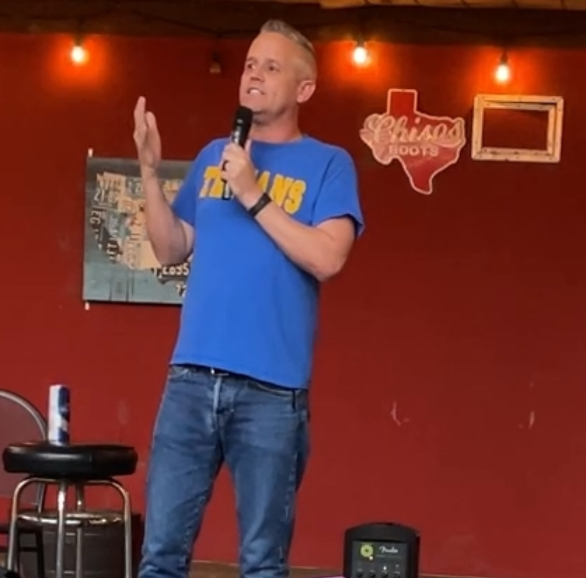 A man in a blue t-shirt and blue jeans holds a microphone and raises his other hand, as though he is in a blues or comedy ...