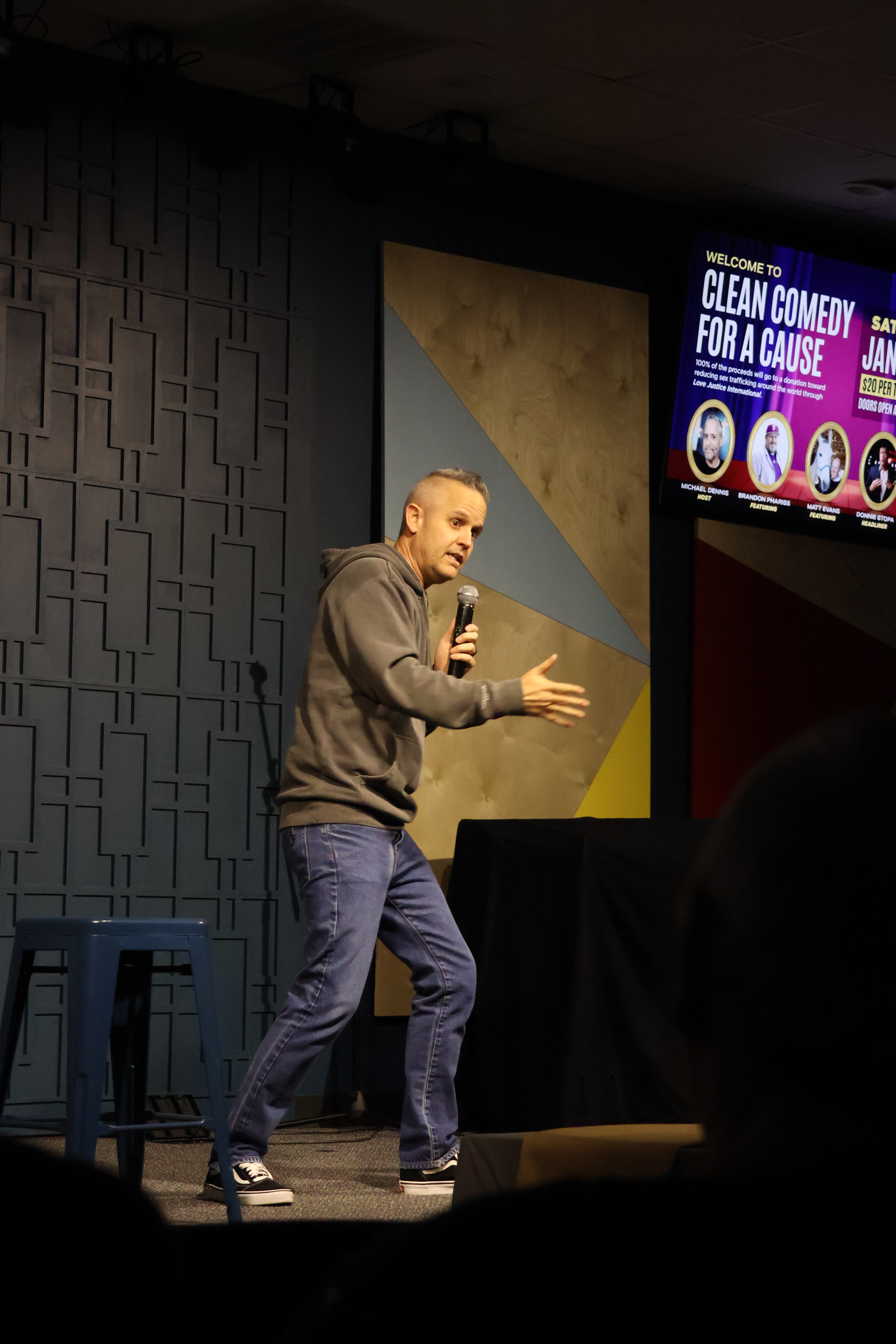 A man in a hoodie and jeans is speaking into a microphone while standing on stage, possibly at a stand-up comedy event. Th...