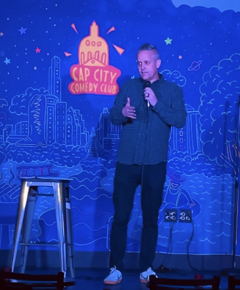 Within an indoor amusement event setting, a man pictured on stage, utilizing a microphone, delivers an entertaining and en...